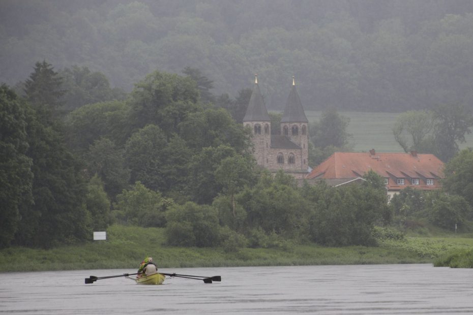Im Hintergrund die beiden Türme des Klosters Bursfelde, davor die Weser und ein Ruderboot im Regen